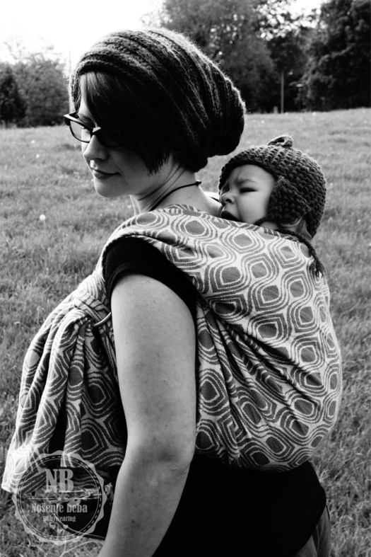 Opis fotografije za slijepe i slabovidne osobe. [Na crnobijeloj fotografiji žena nosi bebu u marami na leđima. U pozadini je livada. Oboje nose ručno pletene kape. Žena zamišljeno gleda prema dolje a beba joj umorno zijeva na leđima. Tkana marama ima uzorak stiliziranog paunovog oka.]